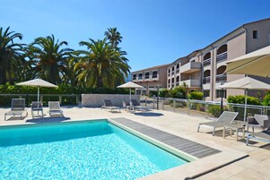 A swimming pool with lounge chairs and umbrellas, surrounded by palm trees and a building with balconies.