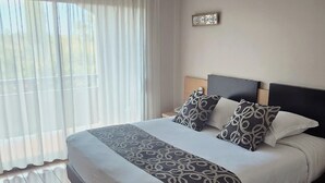 Una cama bien hecha con almohadillas estampadas y un cabecero, una mesita de noche y una ventana con cortinas de encaje.