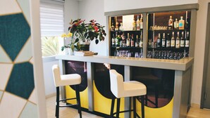 Una zona de bar con dos taburetes blancos, una estantería con botellas y una planta en maceta.