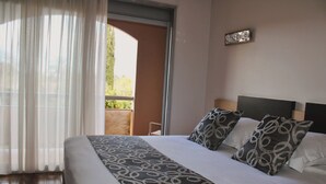 Habitación de hotel con una cama grande, vista a un balcón exterior por una puerta, y una mesa pequeña con una silla.