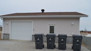 Il s’agit d’un bâtiment d’un seul étage, doté d’une porte de garage et d’une petite fenêtre. Devant le bâtiment, on peut voir quatre poubelles noires alignées sur une allée pavée.