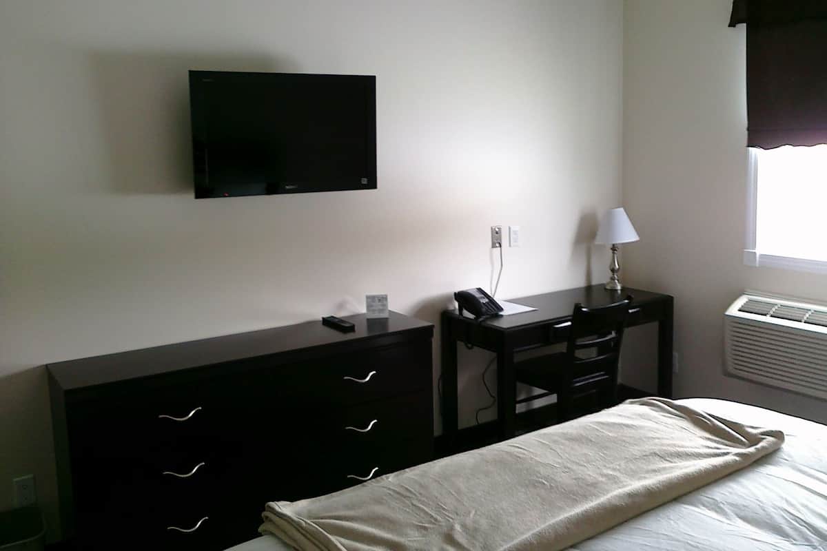 A hotel room with a bed, a desk with a telephone, a flat-screen TV mounted on the wall, and a window with a curtain.