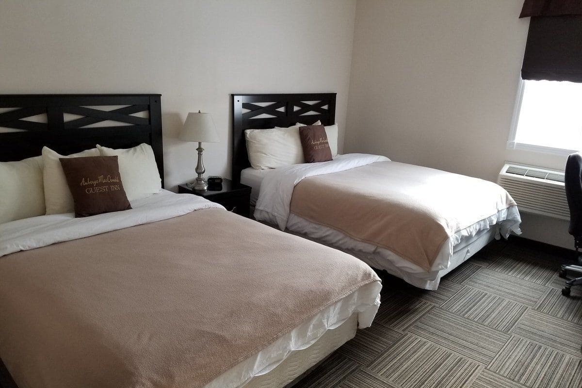 A hotel room with two beds, each with a headboard, a bedside table with a lamp, and a window with curtains.