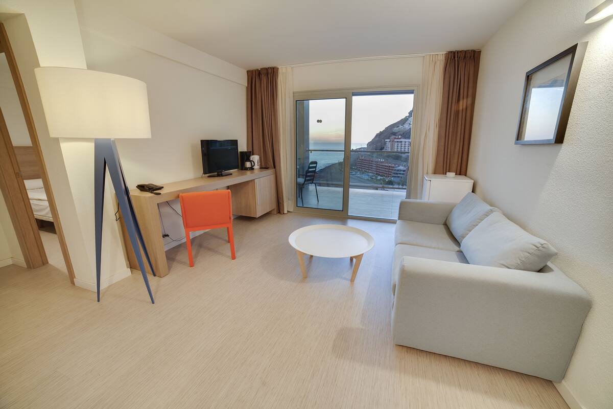 Moderni hotellihuone, jossa on parveke, työpöytä tietokoneella, sohva ja lamppu.
