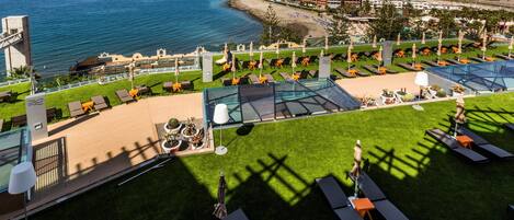 A rooftop pool area with sun loungers and umbrellas, overlooking a beach and ocean.