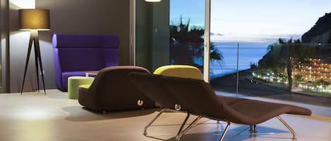 Una sala de estar moderna con un amplio ventanal con vistas a la costa, un sillón morado, un reposapiés amarillo y una lámpara de pie.