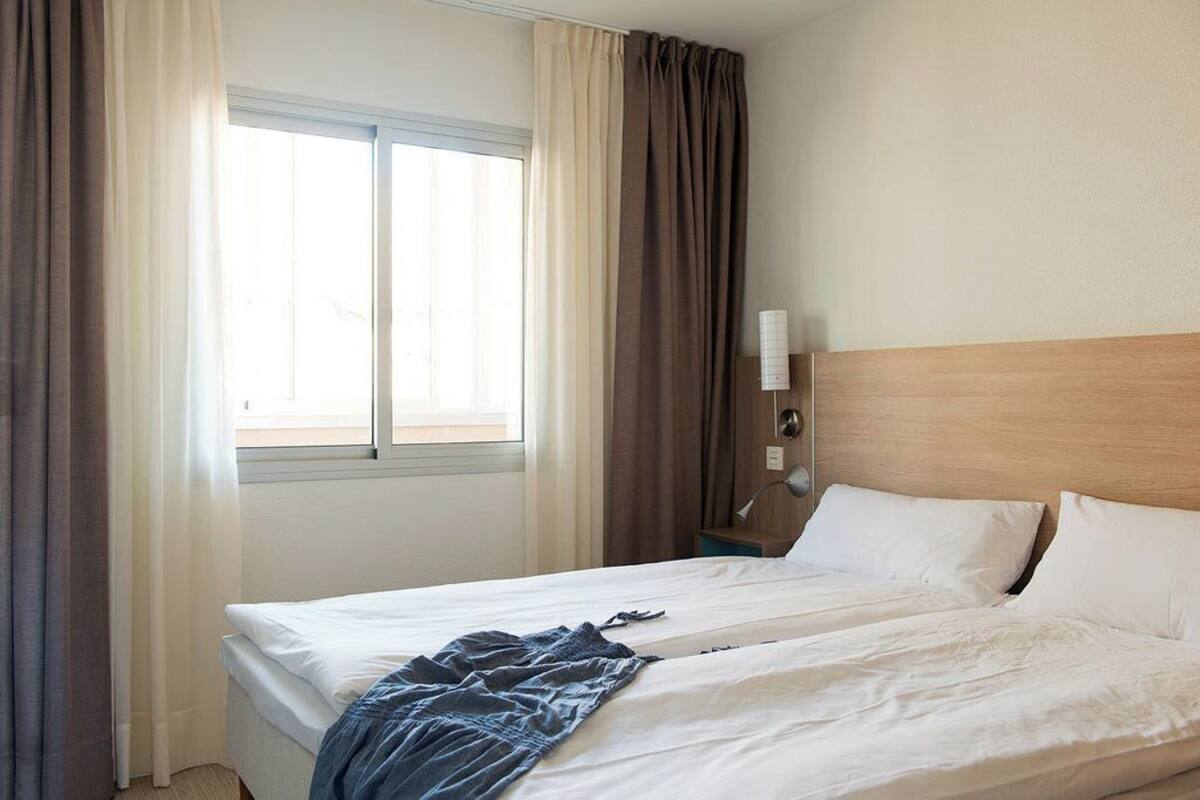 Hotellihuone, jossa on sänky, verhot, ikkuna, yöpöytä ja lamppu.