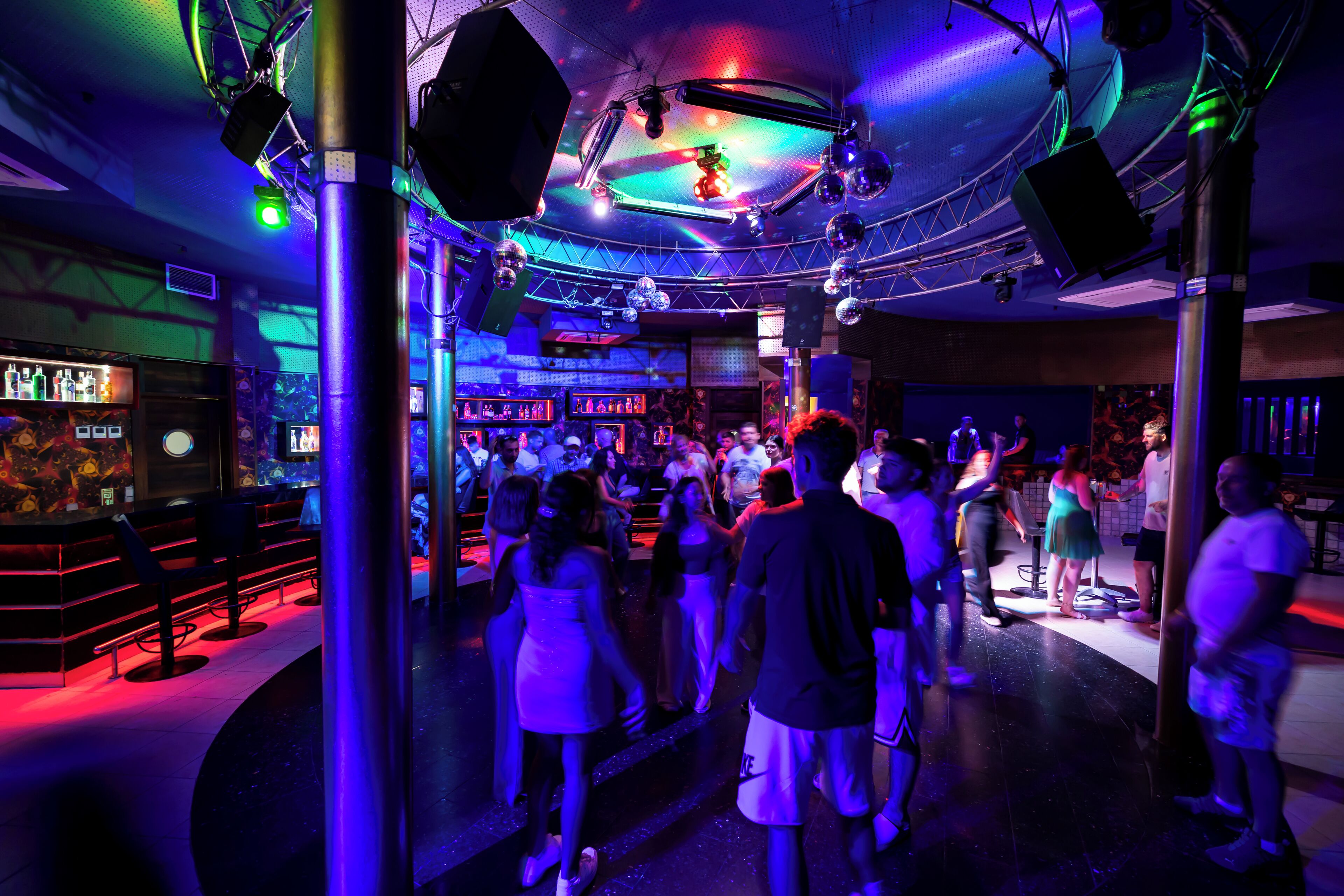 Un lugar interior concurrido que incluye una zona de bar, gente caminando y varios elementos de iluminación.
