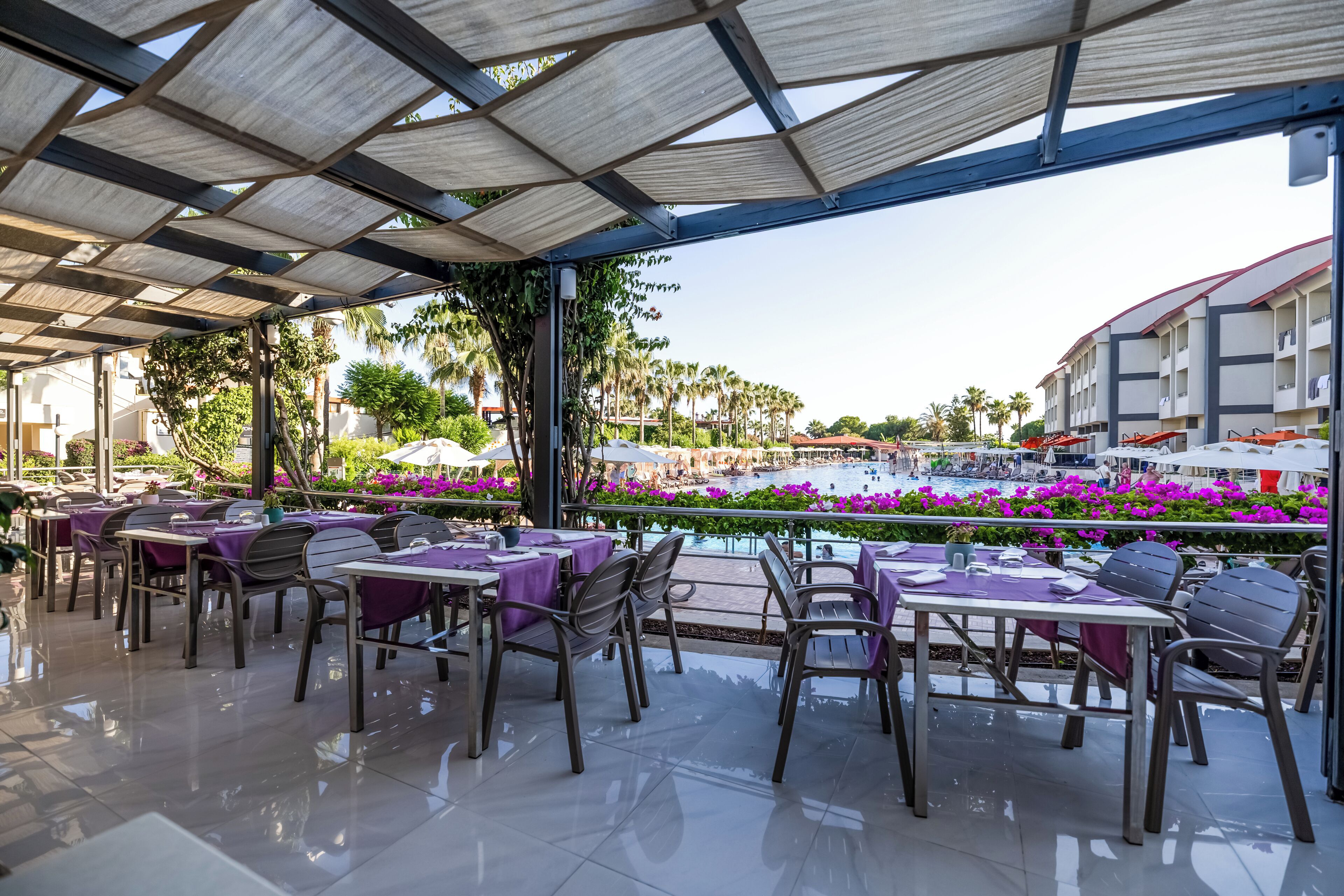 An outdoor dining area with tables and chairs, a pool with purple flowers, and a building in the background.
