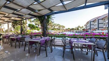 An outdoor dining area with tables and chairs, a pool with purple flowers, and a building in the background.