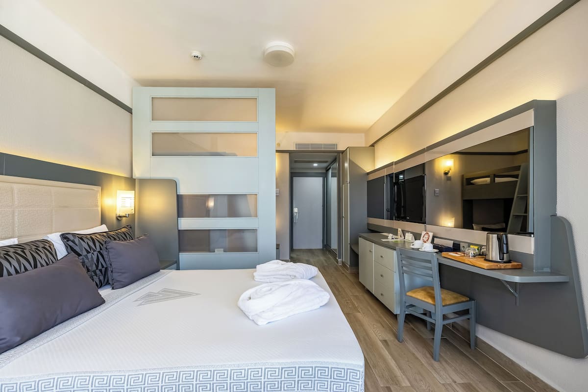 A hotel room with a bed, a desk, a chair, a mirror, and a small kitchenette.