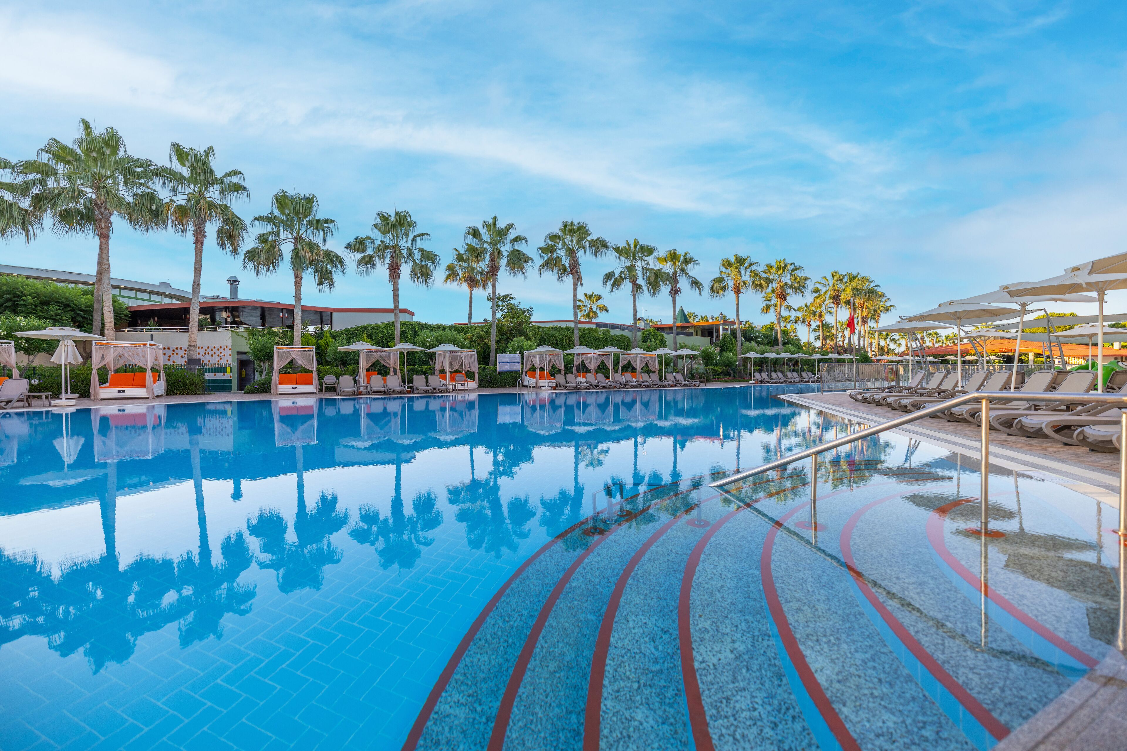 Una piscina con sillones de descanso y sombrillas, rodeada de palmeras y edificios.