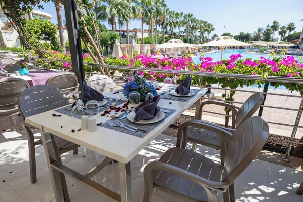 An outdoor dining area with tables set for two, overlooking a pool and surrounded by vibrant flowers.