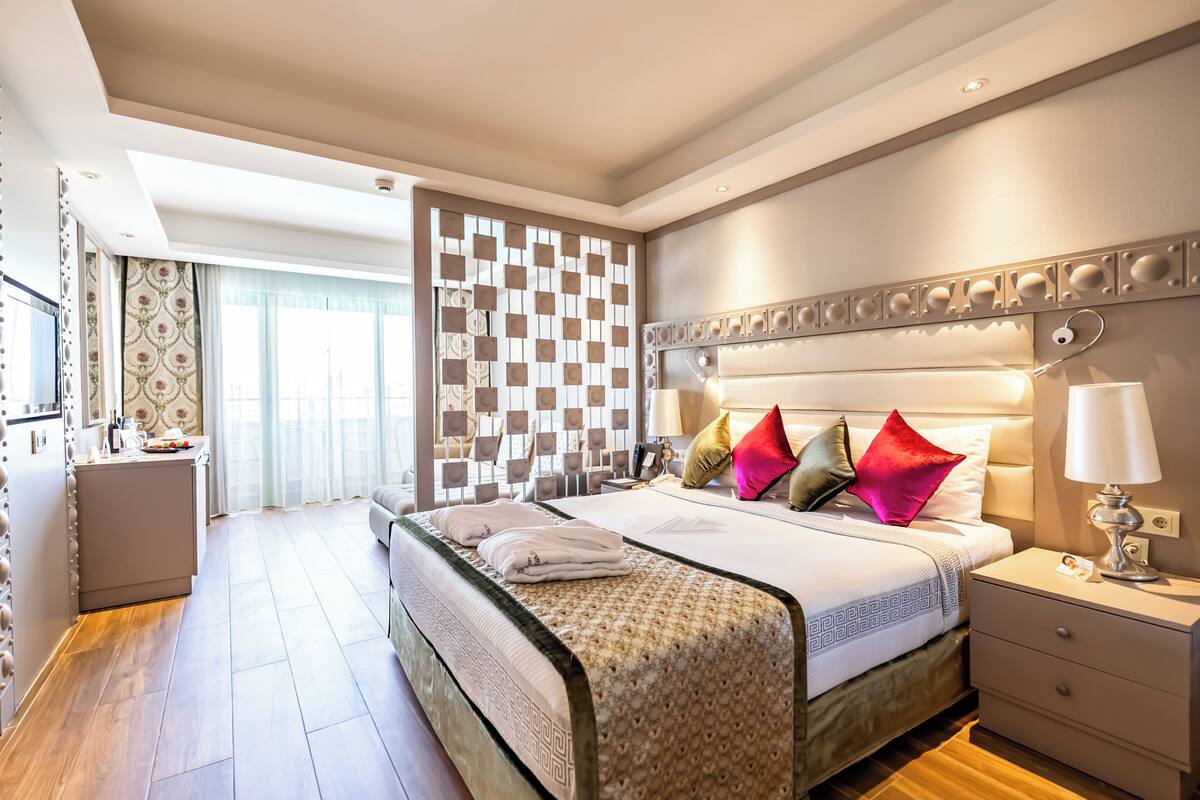 A spacious bedroom with a large bed, a bedside table, a nightstand, and a view of the city through large windows.