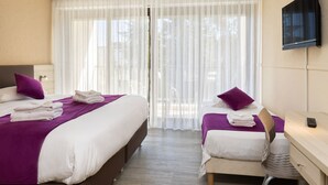 Quarto de hotel com duas camas, uma escrivaninha, uma cadeira, televisão e uma janela grande com cortinas leves.