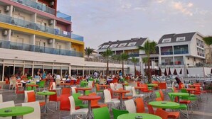 An outdoor seating area with multiple tables and chairs, a multi-story building with balconies, and palm trees in the background. - Side Win Hotel (Manavgat)