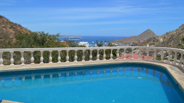 A swimming pool with a white balustrade, surrounded by greenery and overlooking a coastal town with buildings and a mountain in the background. - Casa Rosa (Cabo San Lucas)