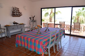 A room with a large window, a table with a colorful tablecloth, a desk with a ship decoration, and a chair.