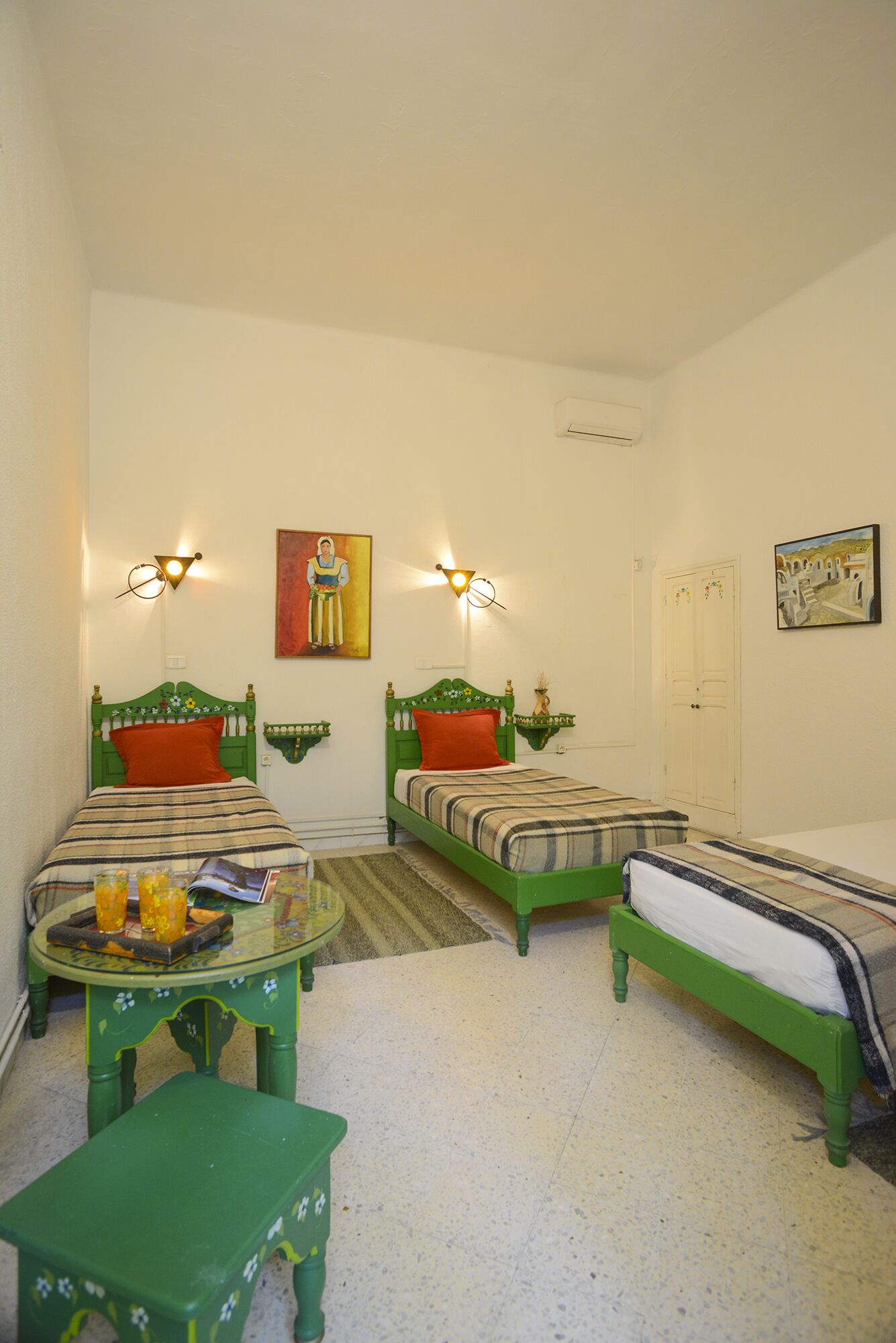 Una habitación con dos camas verdes, cada una con una almohada roja, una mesita redonda con superficie de vidrio y un taburete verde. Hay lámparas de pared y un cuadro sobre las camas.