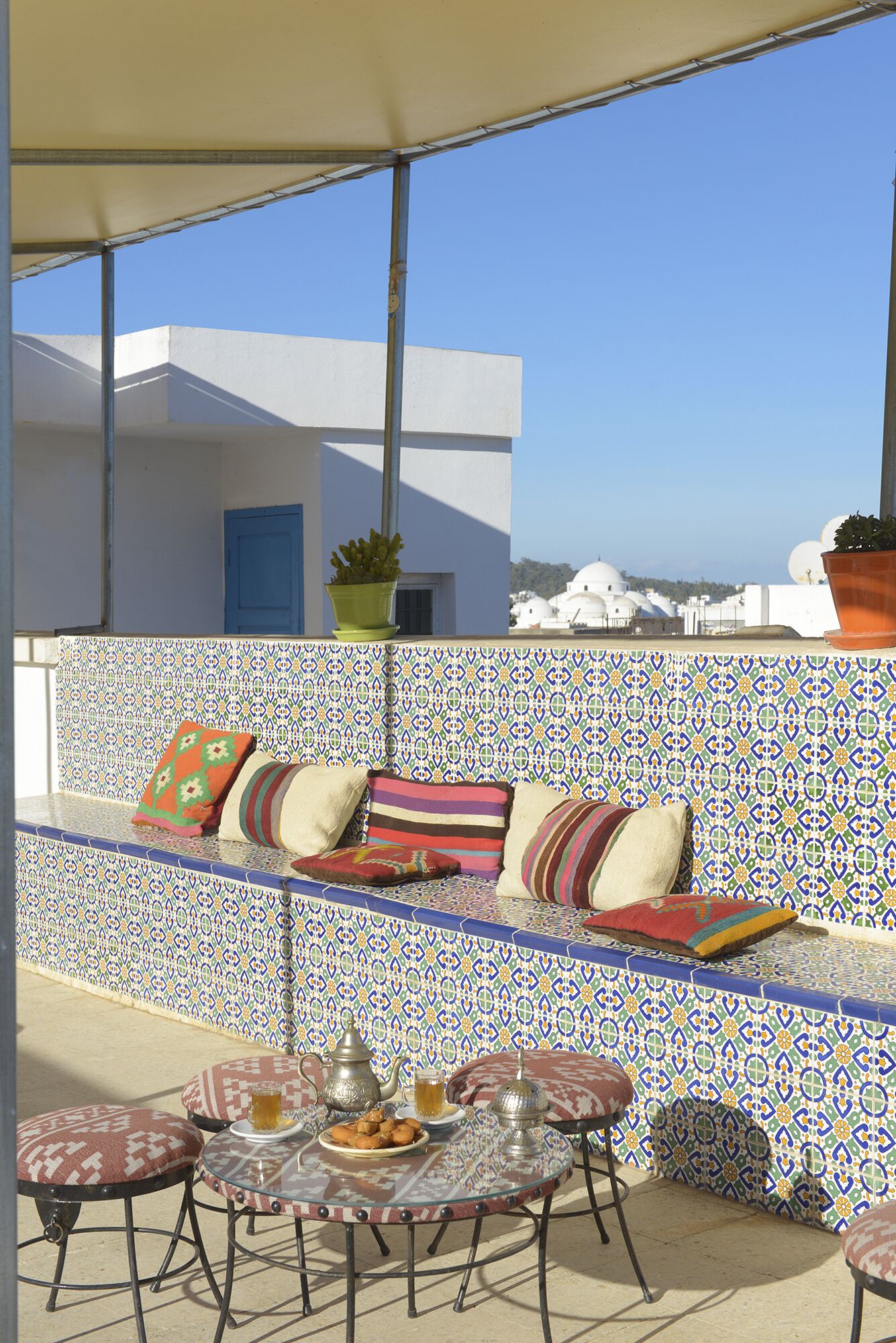 A rooftop terrace with a patterned wall, a seating area with cushions, a table with a teapot and snacks, and a view of buildings in the background.