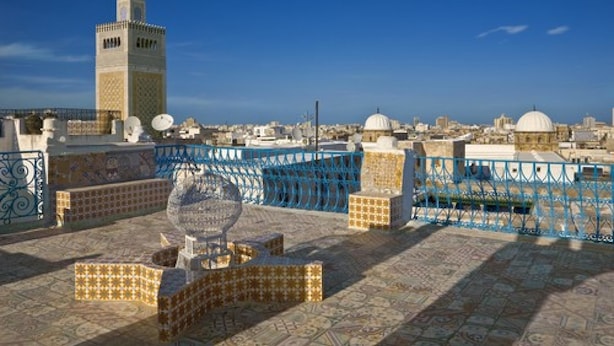 A rooftop terrace with patterned tiles, a decorative glass sculpture, and a view of a cityscape including a prominent tower. - Dar Ya - Hostel (Tunis)