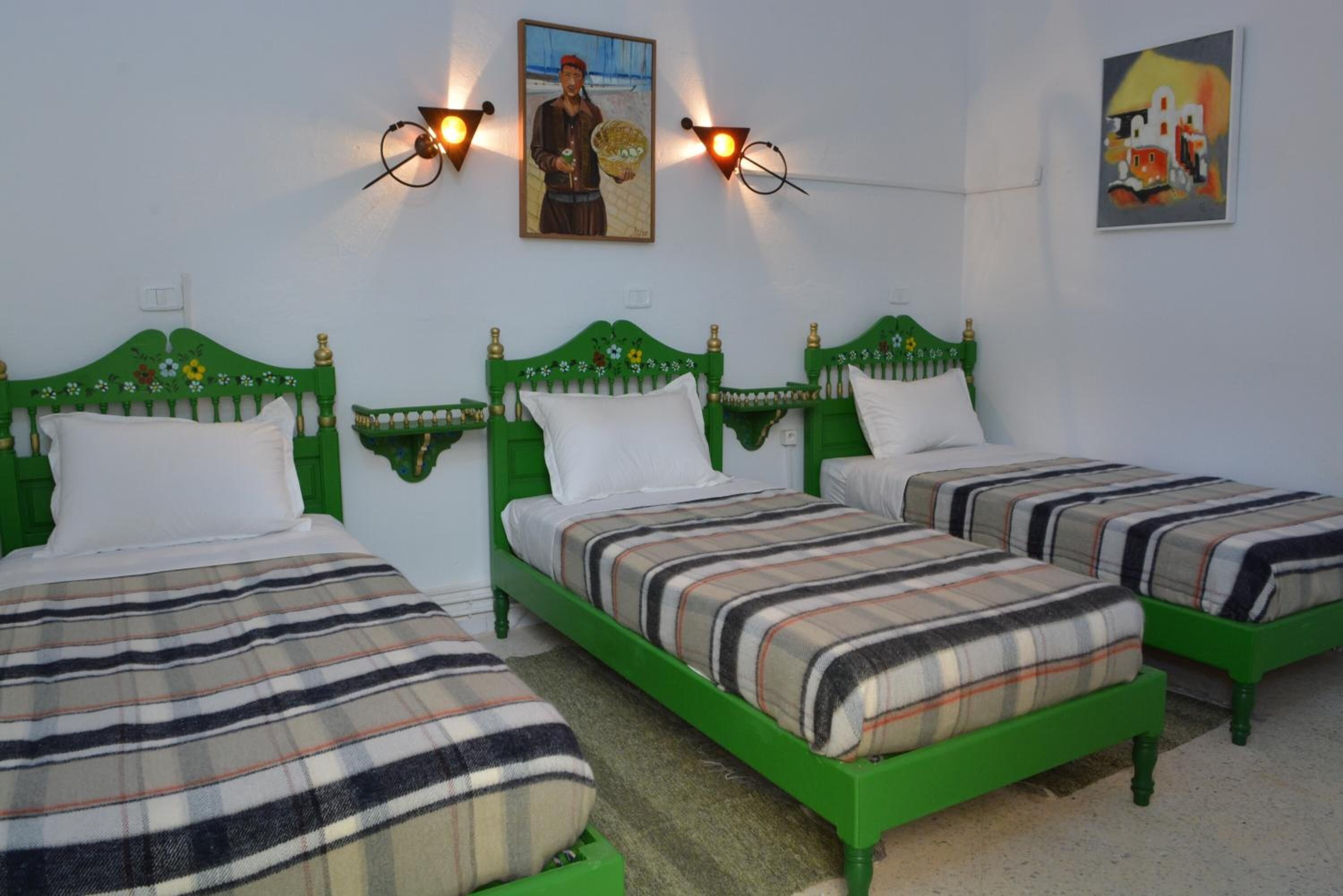 Une chambre avec trois lits verts, des oreillers blancs et des couvertures à rayures. Des lampes sont fixées aux murs et un tableau est accroché au-dessus des lits.