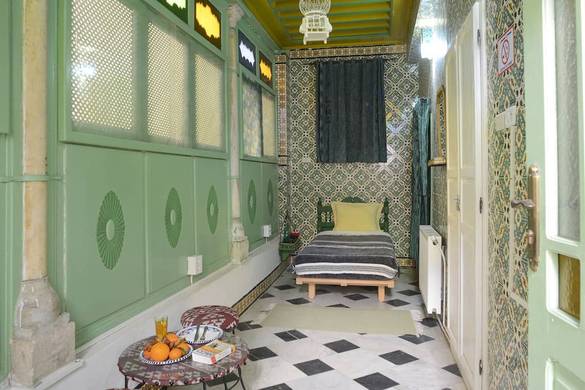 A room with a checkered floor, a bed with a striped blanket, a table with oranges and a book, and a fan on the ceiling.