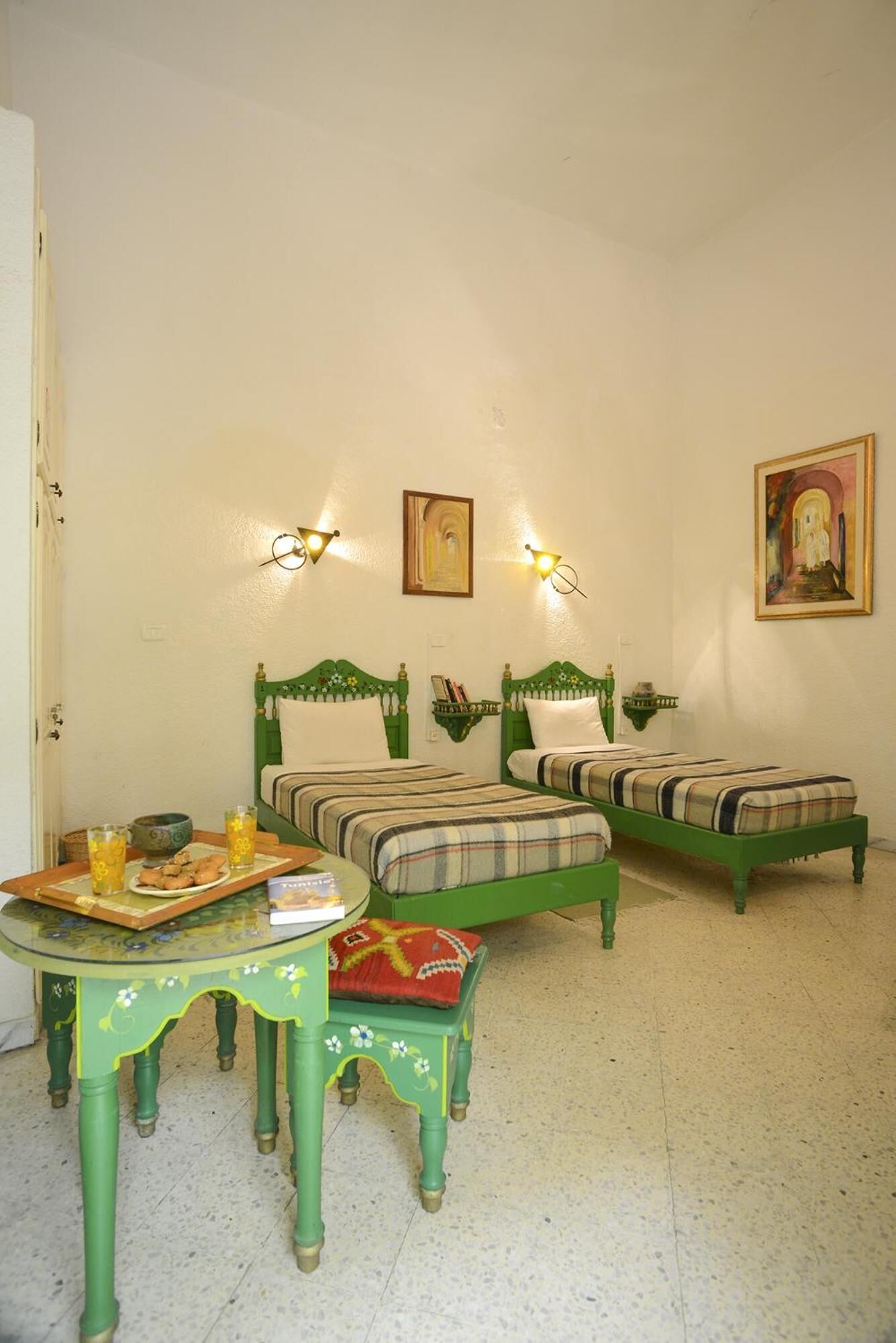 Una habitación con dos camas verdes, una mesita con superficie de vidrio y un estante de pared con libros y un cuenco.