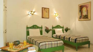Una habitación con dos camas verdes, una mesita con superficie de vidrio y un estante de pared con libros y un cuenco.