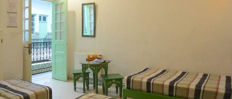 A room with two beds, a small table, and a chair. There is a door leading to an outdoor area.