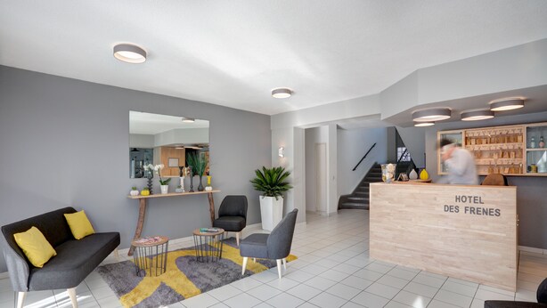 Un área de recepción de hotel moderna con una paleta de colores gris y blanco, un mostrador de recepción con la leyenda “HOTEL DES FRENES” y una escalera que conduce al piso superior.