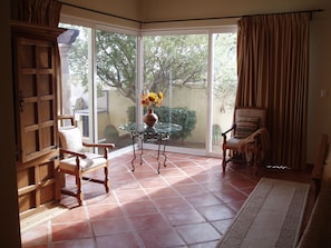 A room with a glass door leading to a garden, a wooden chair, a glass table with a vase, and a patterned rug.
