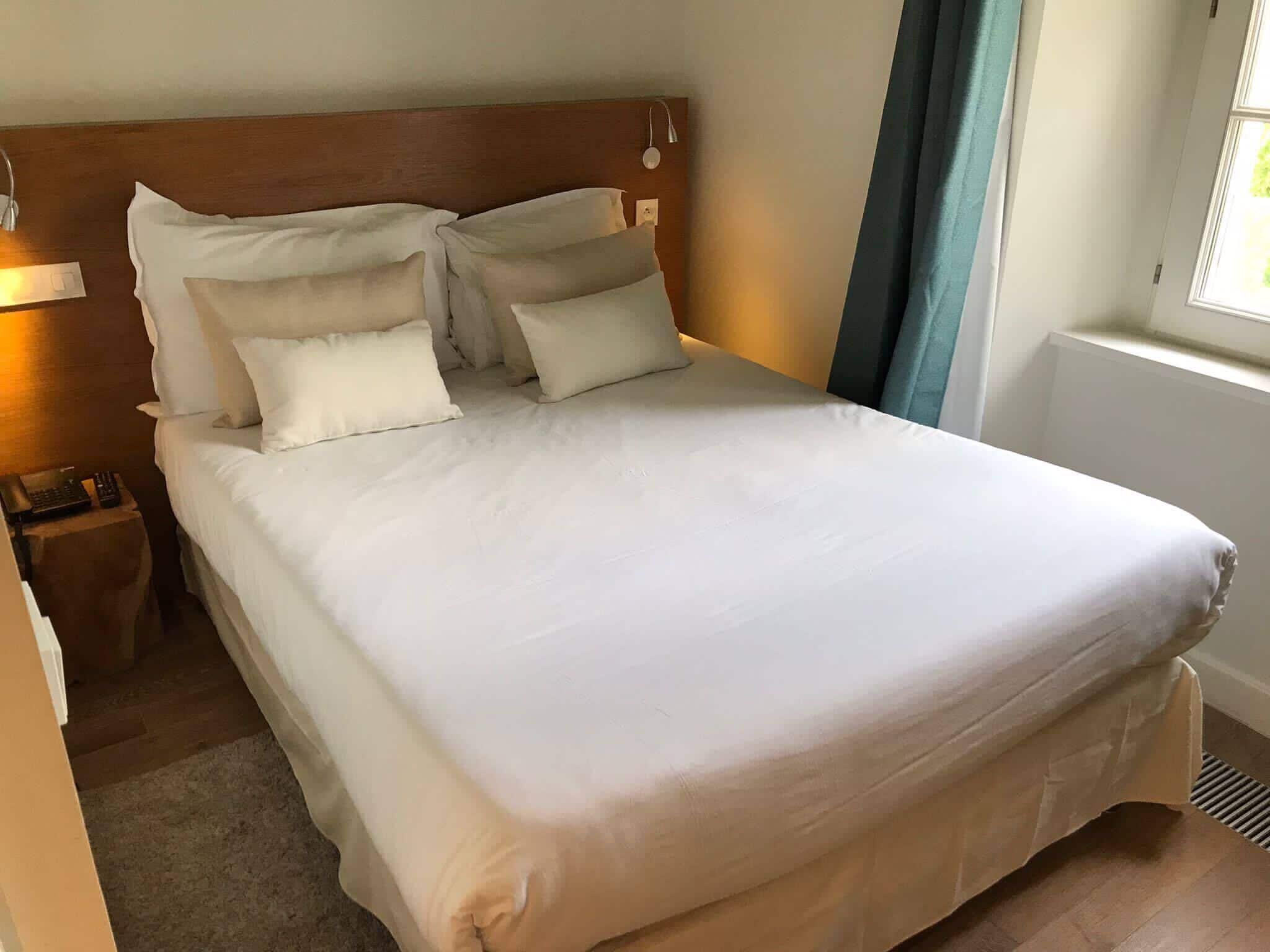 Een net opgemaakt bed met witte lakens en meerdere kussens, een houten hoofdbord, een nachtkastje met een lamp en een raam met gordijnen.