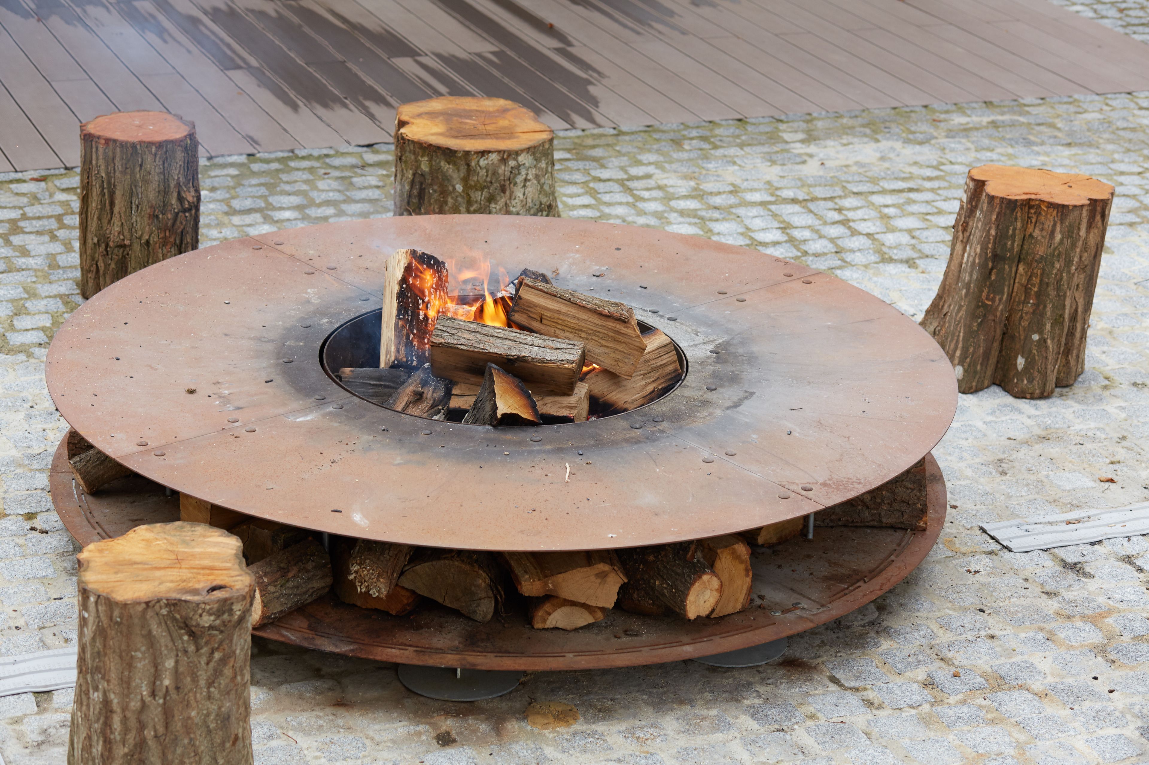 A circular fire pit with logs and flames, surrounded by four wooden stumps, set on a cobblestone surface.