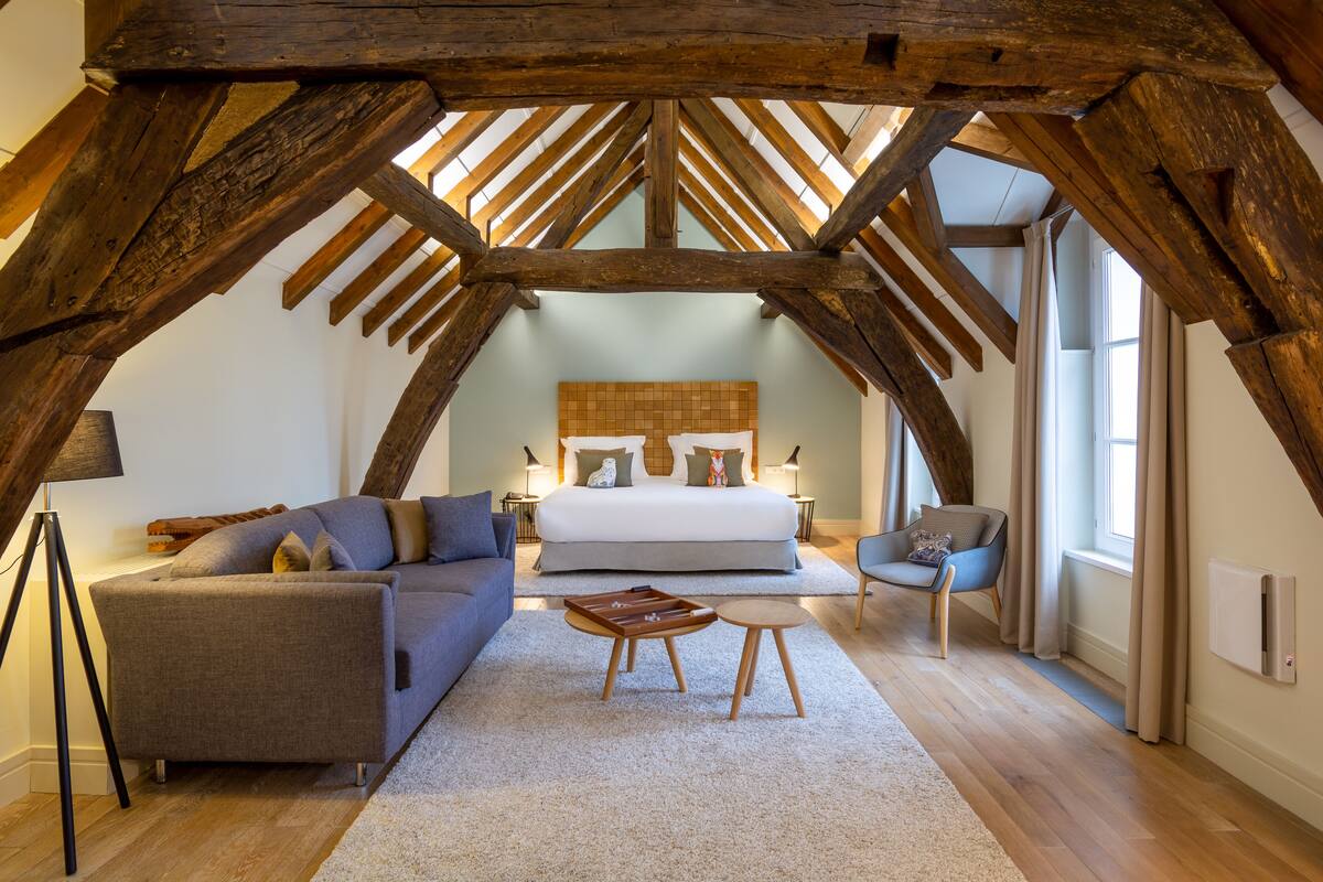 A bedroom with a large bed, a sofa, a small table, and a chair. The room has wooden beams on the ceiling and a window with curtains.