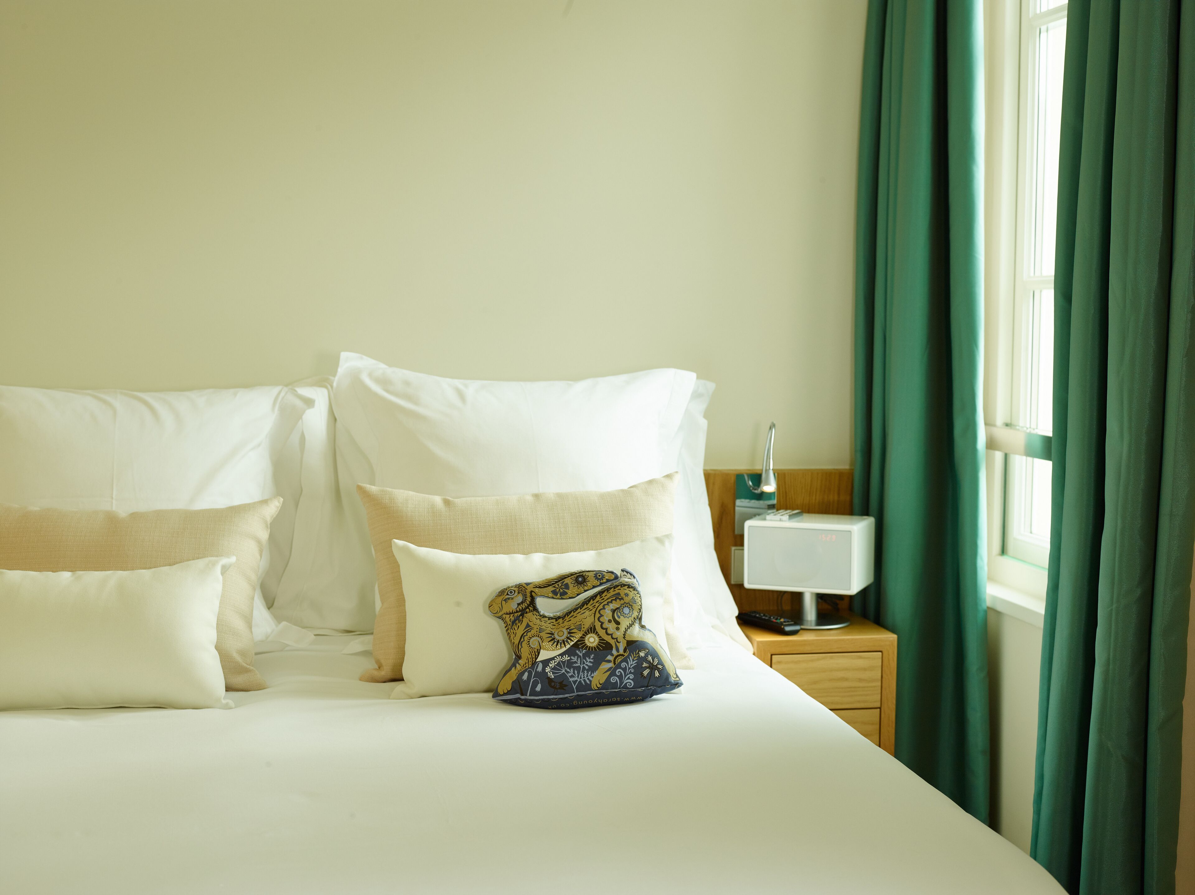 Una cama bien hecha con almohadas blancas y beige, una almohada decorativa con estampado floral, una mesita de noche con lámpara y una ventana con cortinas verdes.
