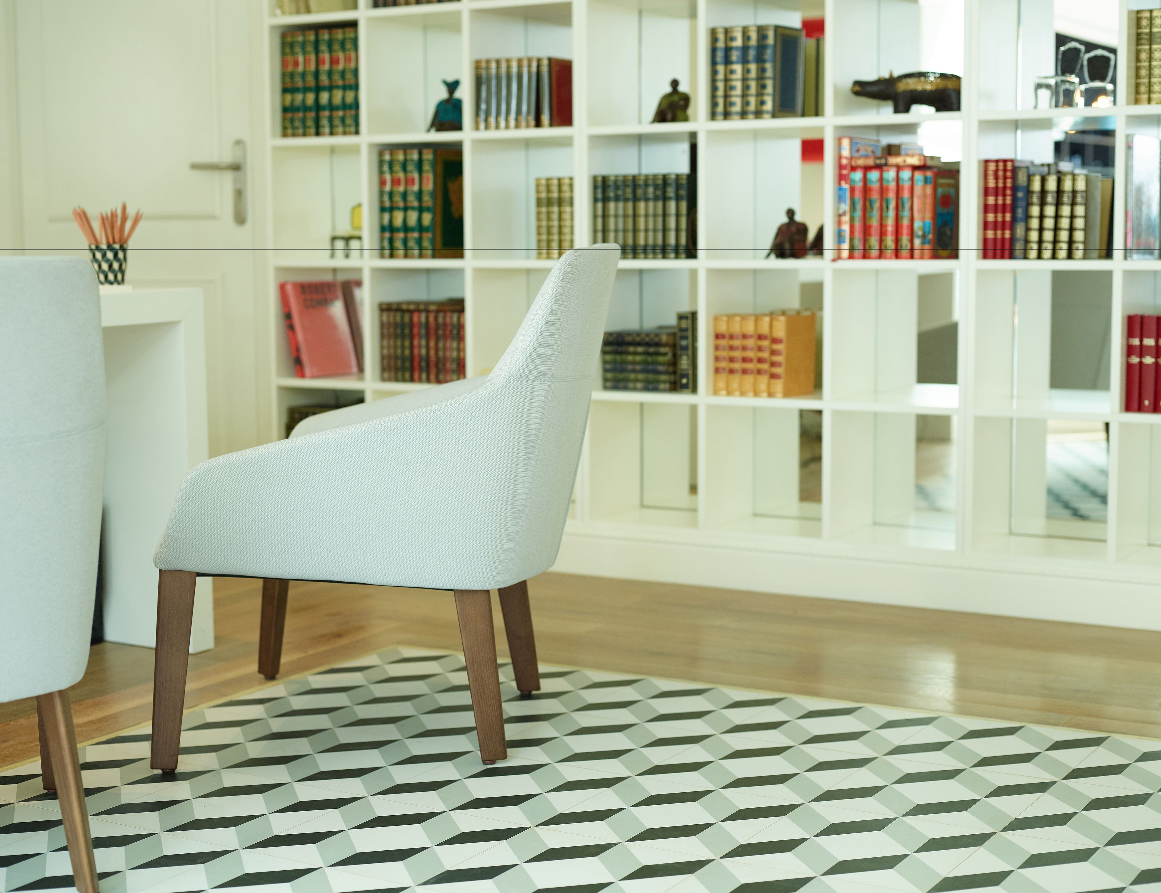 Habitació moderna amb un paviment de disseny geomètric, una cadira tapissada de blanc i una estanteria plena de llibres i objectes decoratius.