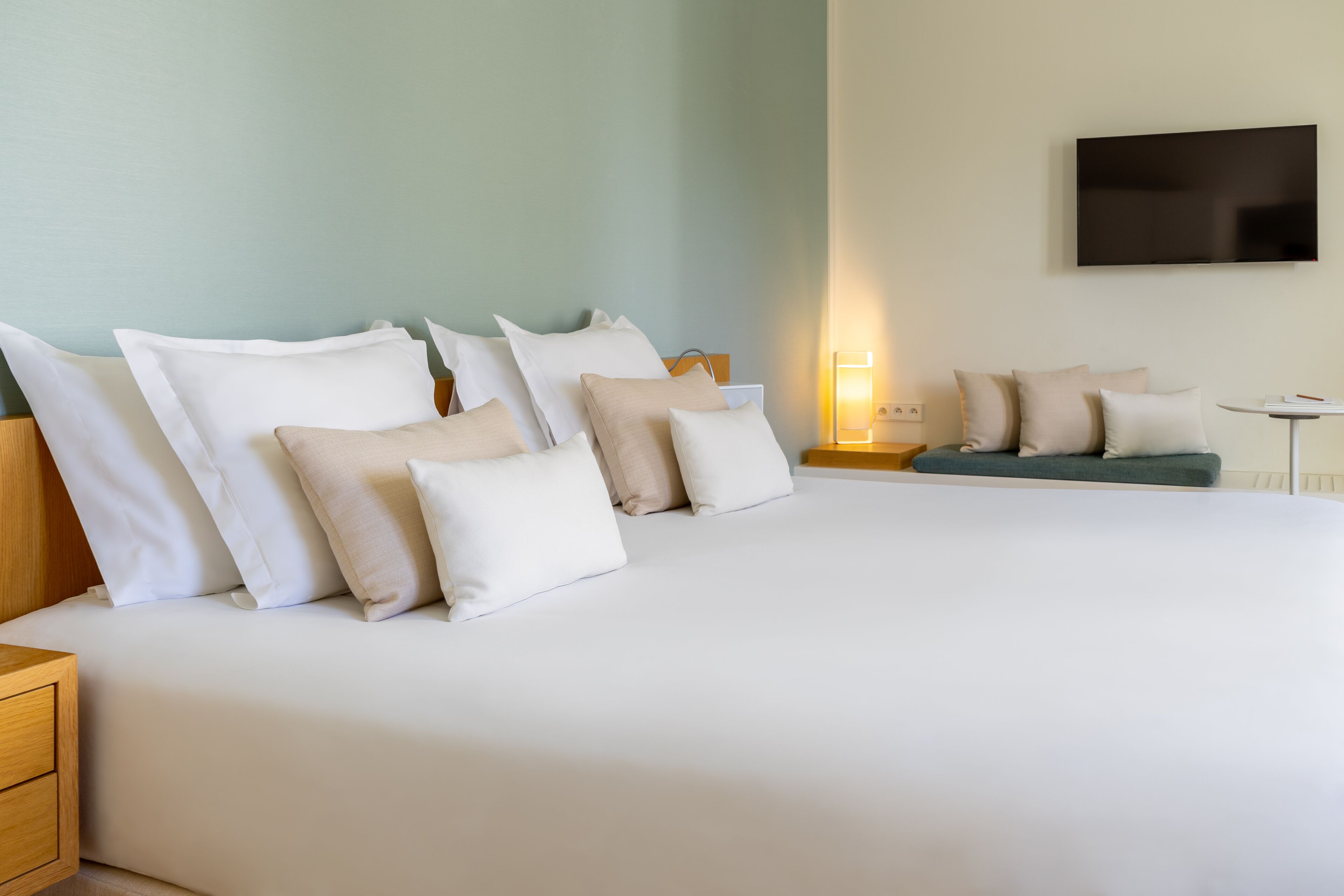 A hotel room with a large bed, multiple pillows, a bedside table, a small table, and a wall-mounted TV.