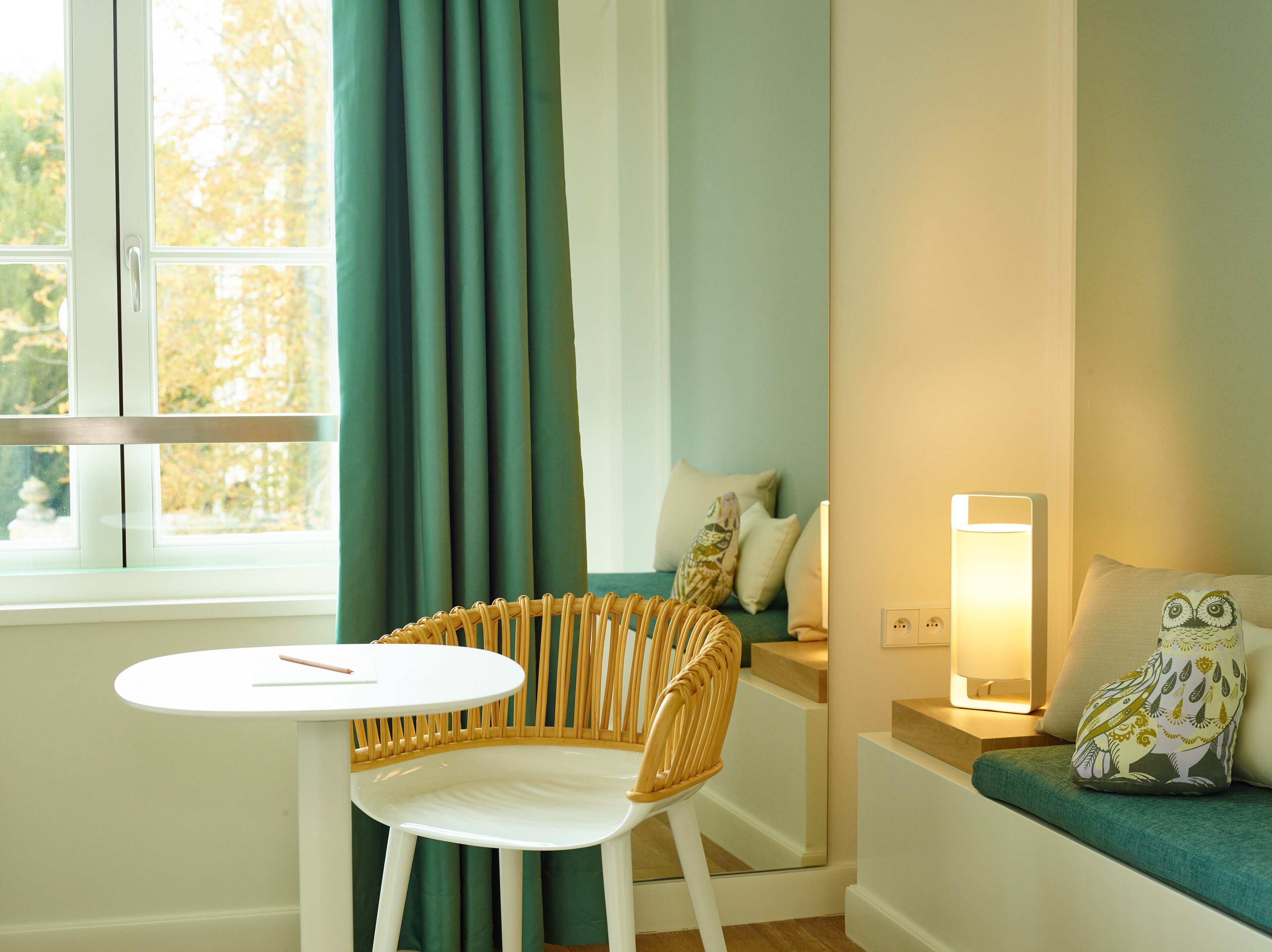 A room with a white chair, a round table, a sofa with a patterned pillow, and a standing lamp.