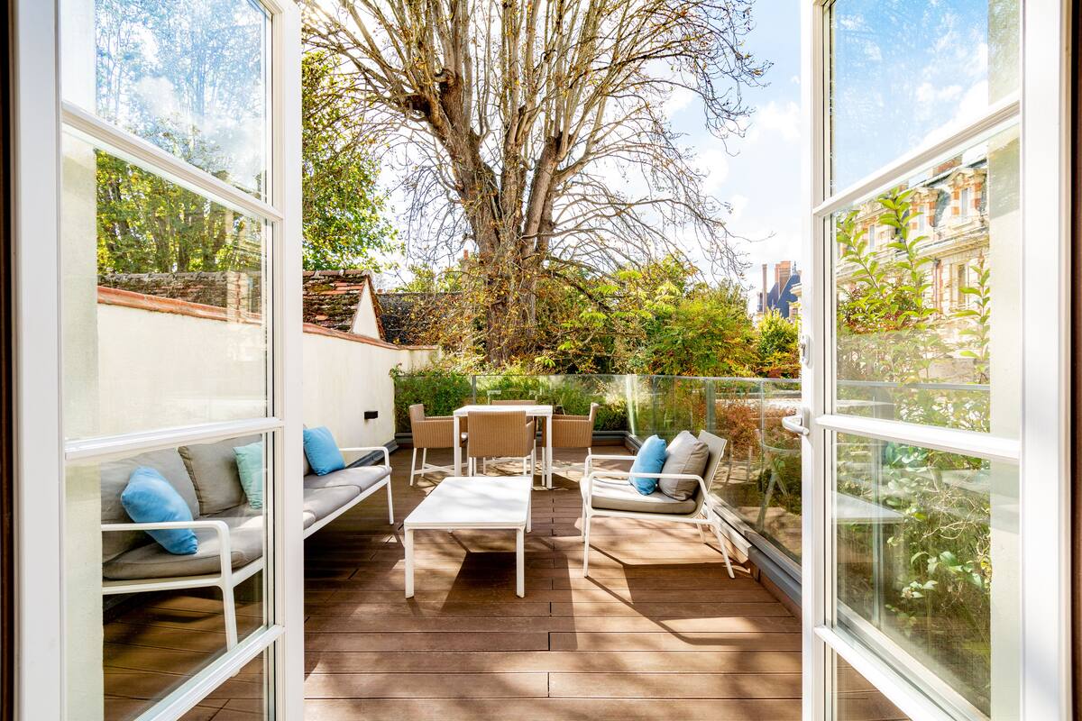 An outdoor patio with a wooden deck, white furniture, and a view of a tree and garden.