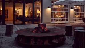 A circular fire pit with logs, surrounded by wooden stools, in front of a building with large windows and an outdoor seating area. - Hôtel de Cavoye (Fontainebleau)