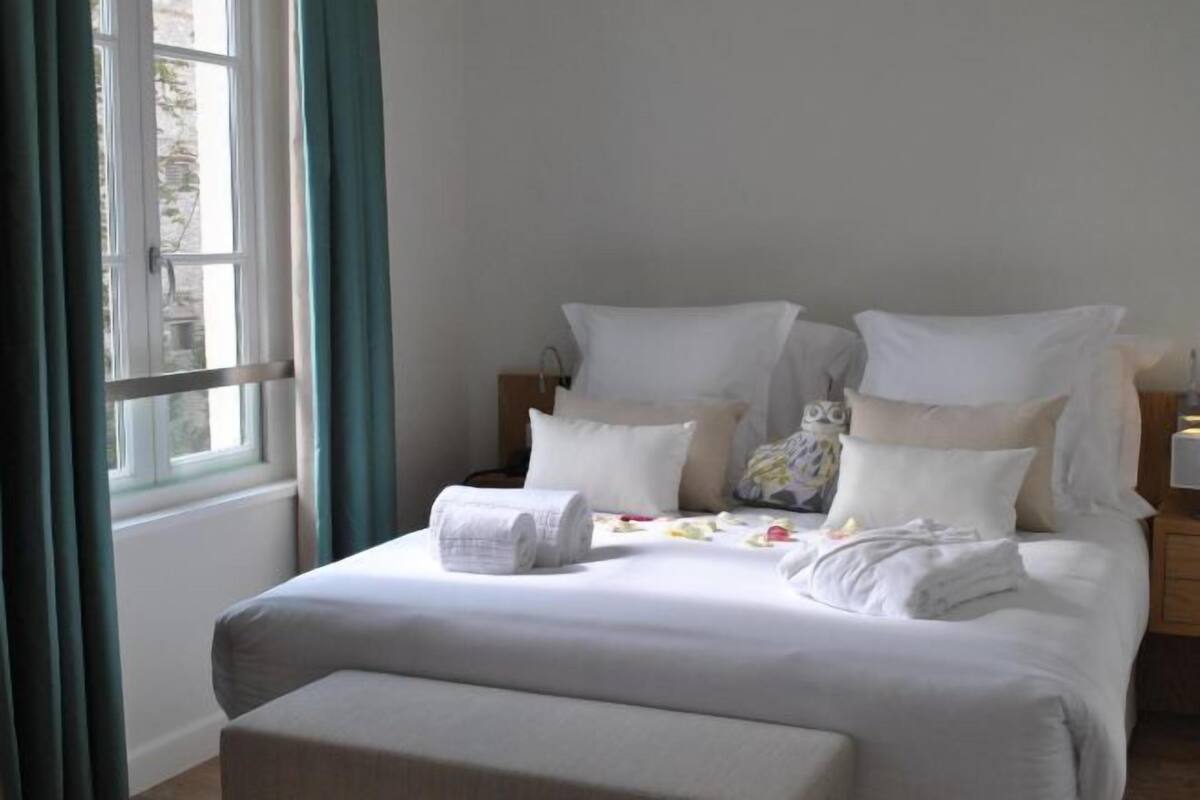 A neatly made bed with pillows and a rolled towel, a bedside table with a lamp, and a window with curtains.