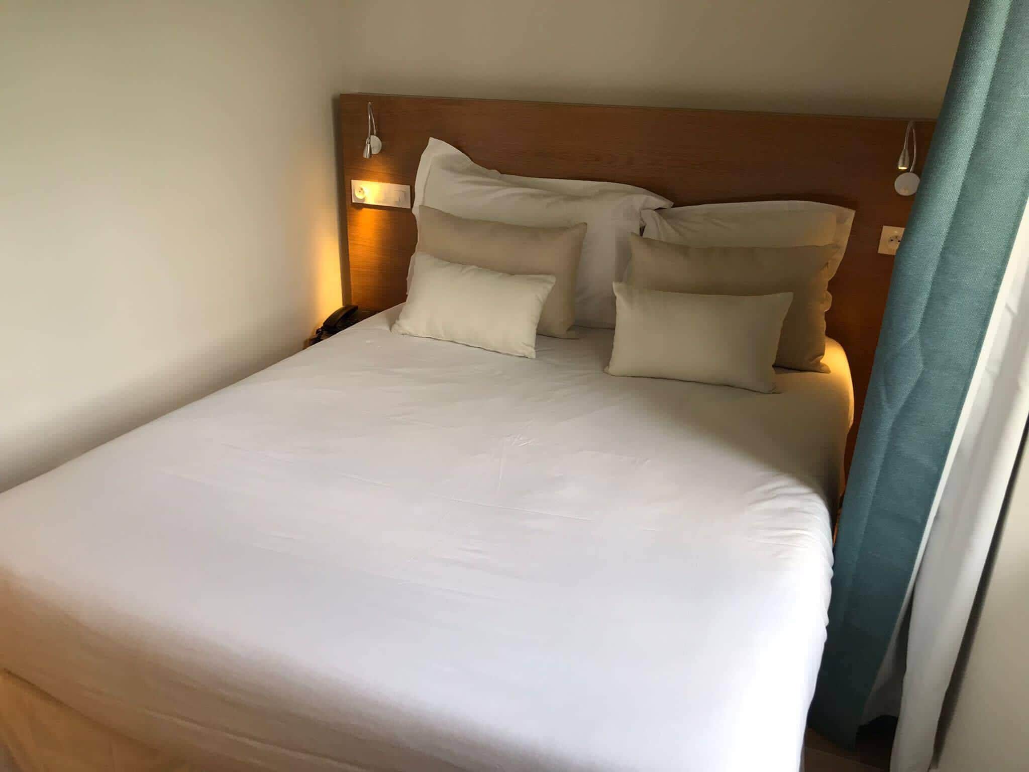 Una cama bien tendida con sábanas blancas y varios almohadones, un cabecero de madera y una lámpara de noche.