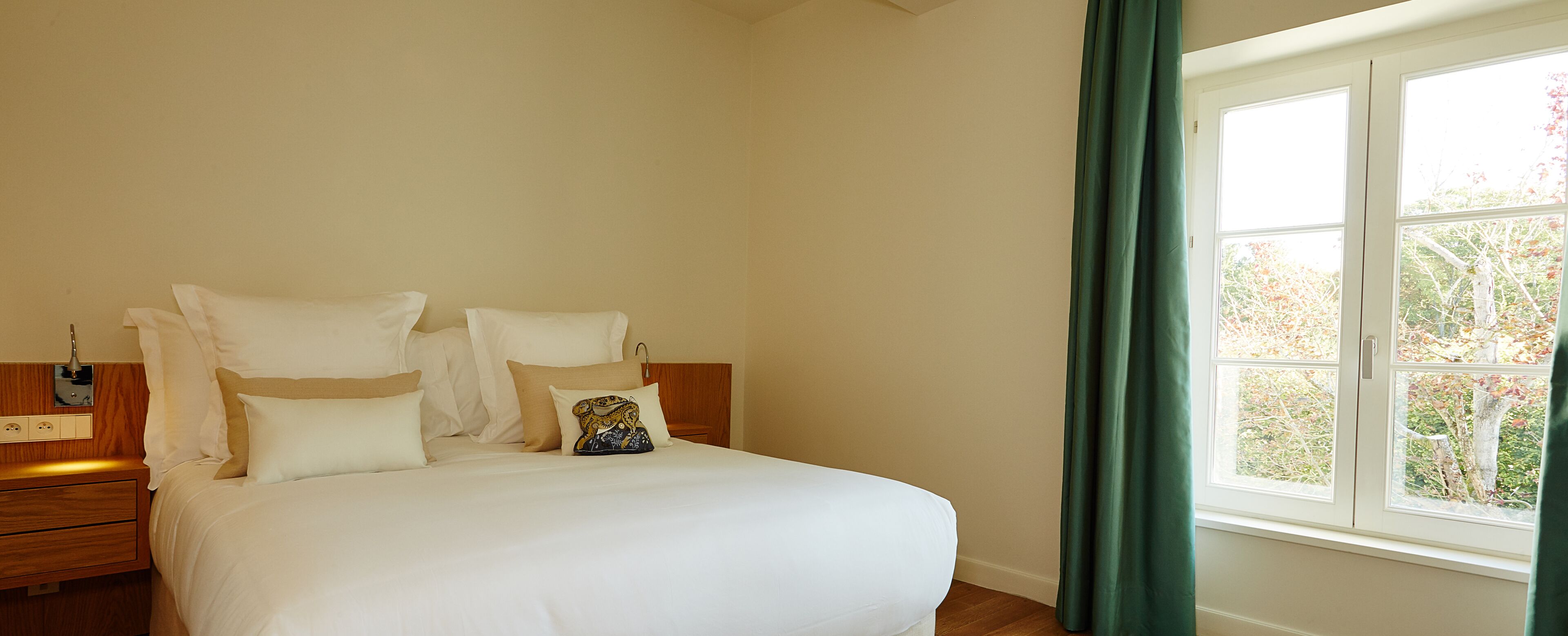 A neatly made bed with white linens and multiple pillows, a wooden nightstand with a lamp, and a window with a view of trees.