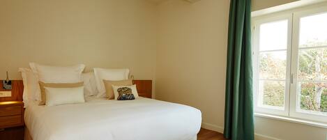 A neatly made bed with white linens and multiple pillows, a wooden nightstand with a lamp, and a window with a view of trees.