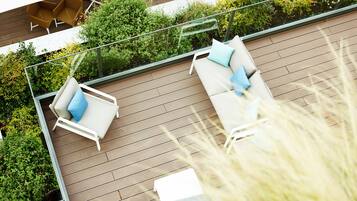 Eine Dachterrasse mit zwei weißen Liegestühlen, einem Glasgeländer und üppiger Bepflanzung.