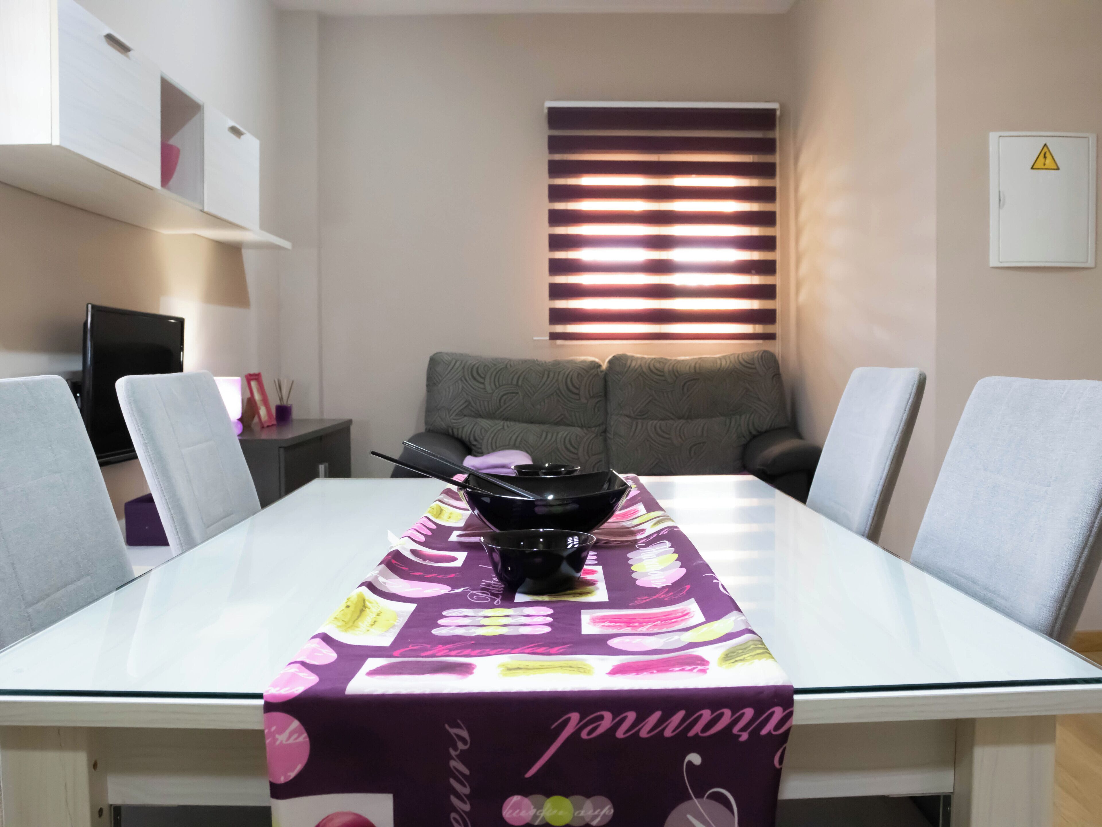 Uma sala de jantar com uma mesa de vidro, toalha de mesa roxa e uma tigela preta. Há cadeiras cinzas, um sofá e uma TV sobre um suporte.