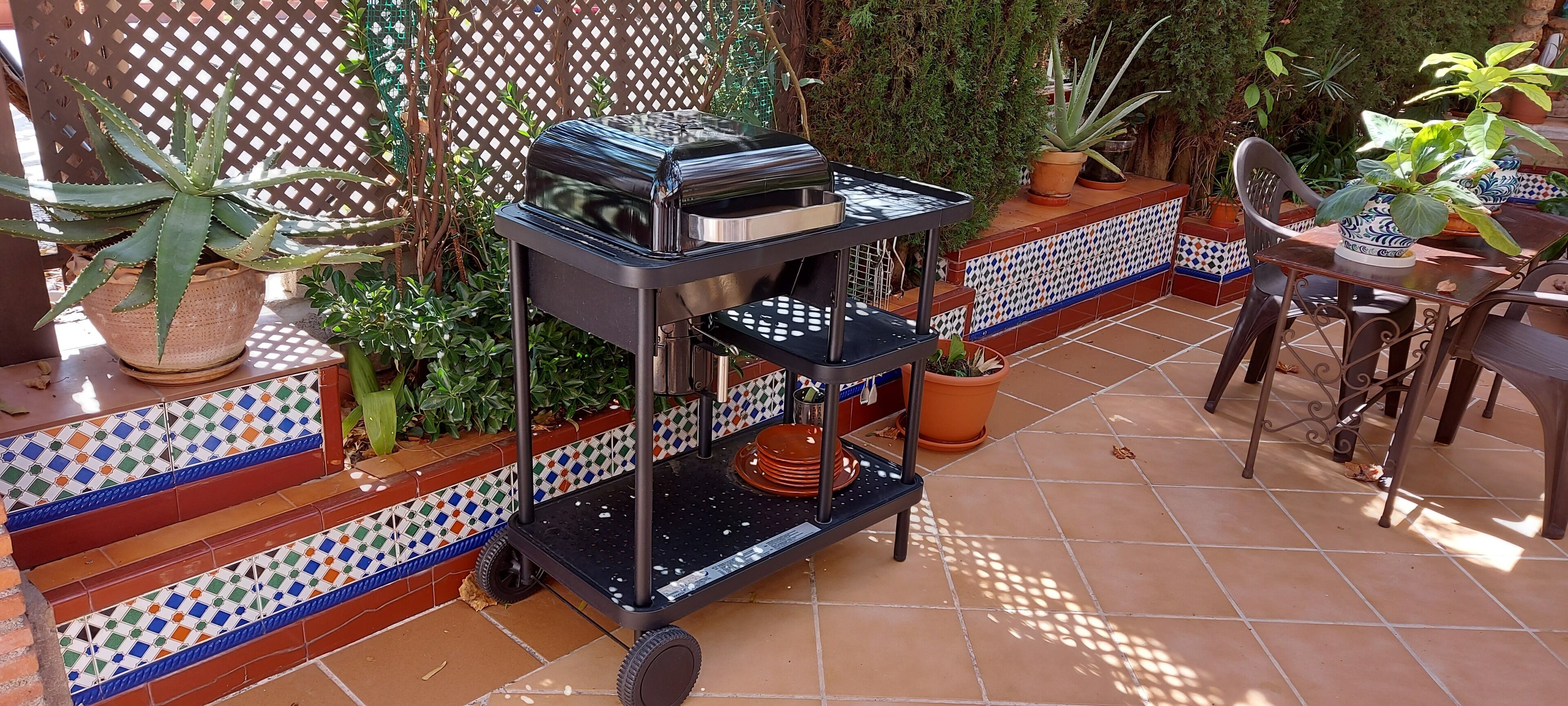 A patio with a grill cart, potted plants, and a tiled bench area.