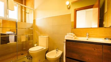 Un baño moderno con mampara de vidrio, inodoro, bidé y tocador de madera con lavabo.
