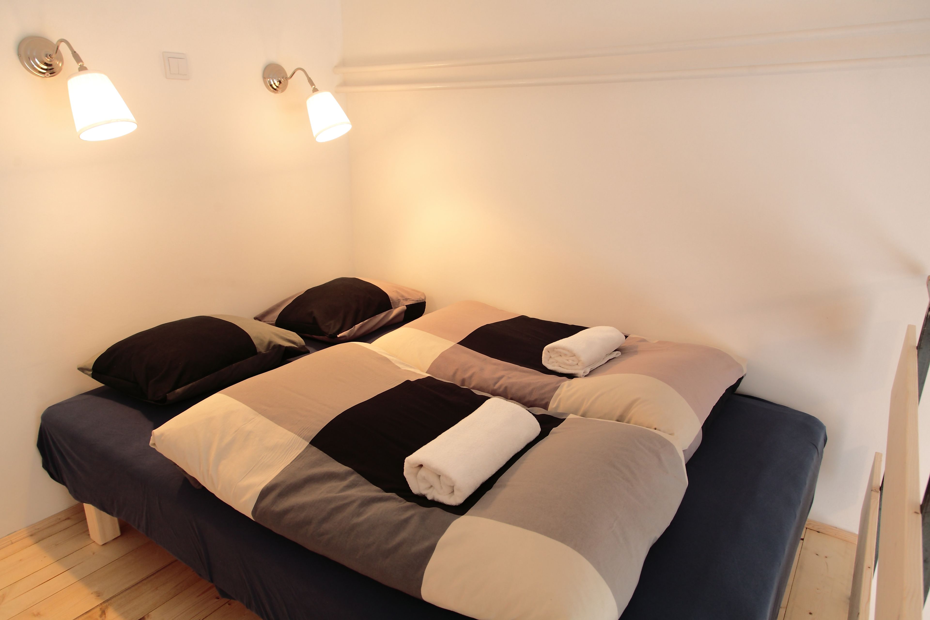 Una cama doble con colcha azul oscuro y color crema, dos lámparas de pared y suelo de madera.